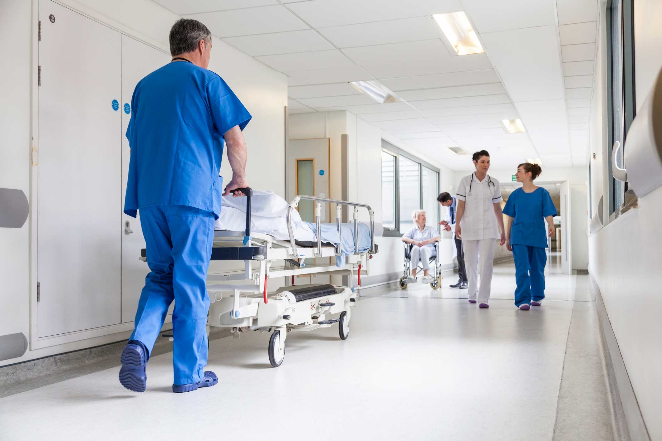 Hospital hallway with man walking bed down hall and two physicians